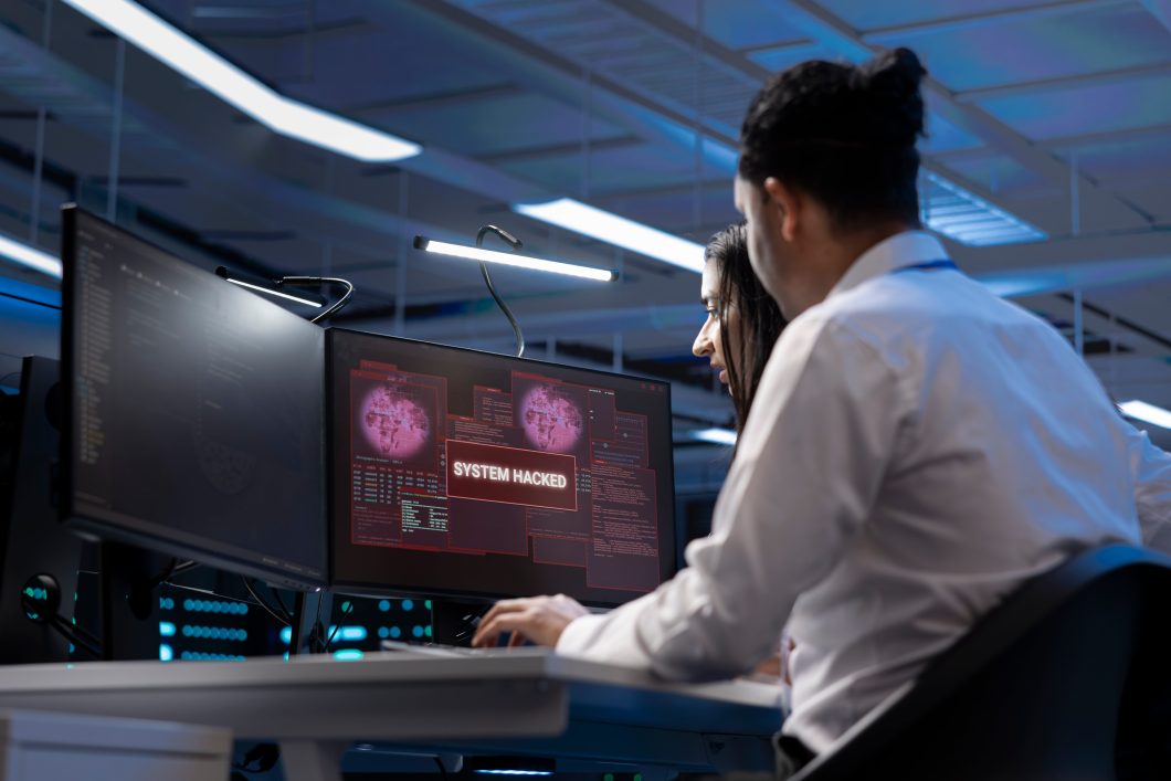 Two people at a computer facing a computer that says it's hacked.